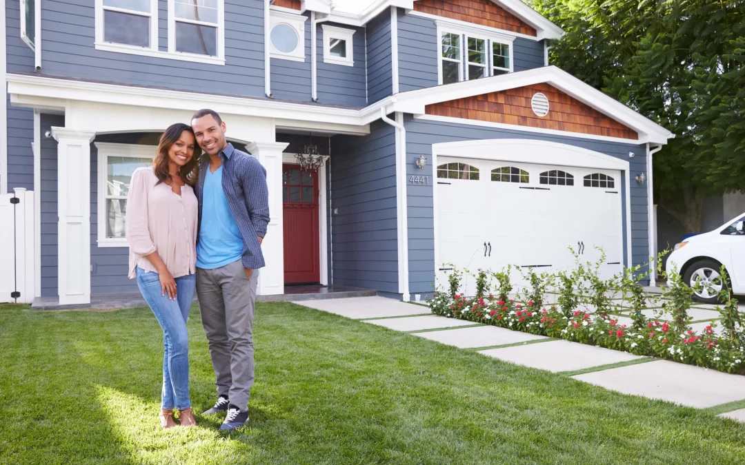 A smiling couple stands in front of a modern, two-story blue house with white trim. The manicured lawn and flowers create a welcoming atmosphere.
