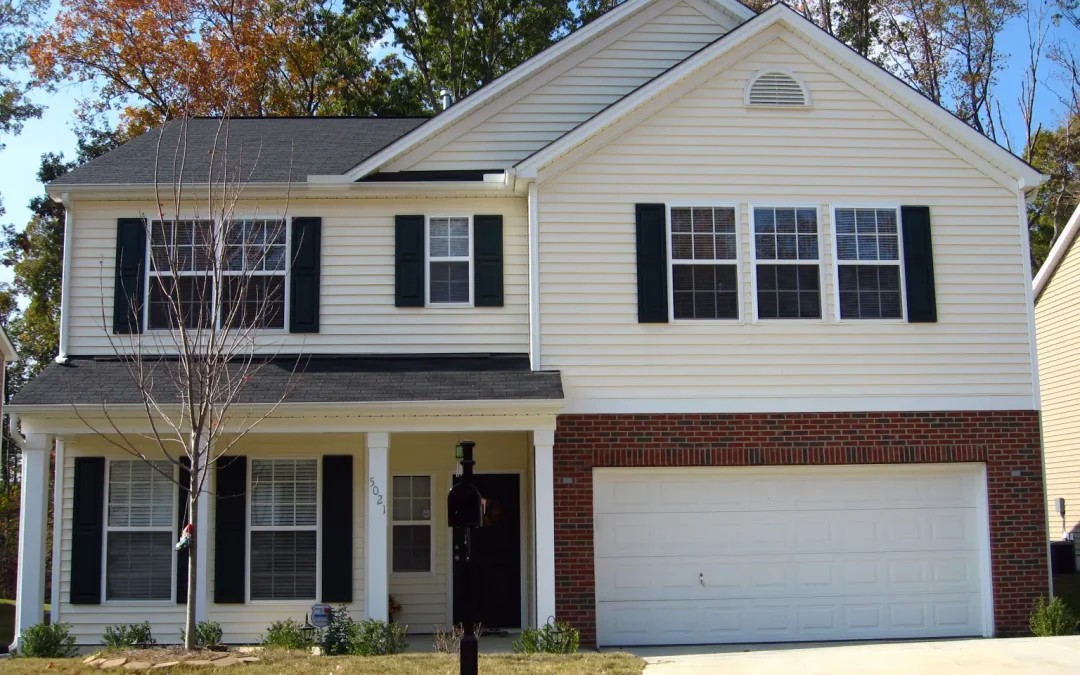 Two-story suburban house with a mix of brick and siding façade, black shutters, and a two-car garage. Tree and mailbox in front yard, sunny day.